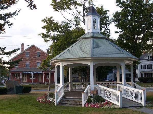 The gazebo on the Sutton town common, Massachusetts, with flower beds and a brick building behind